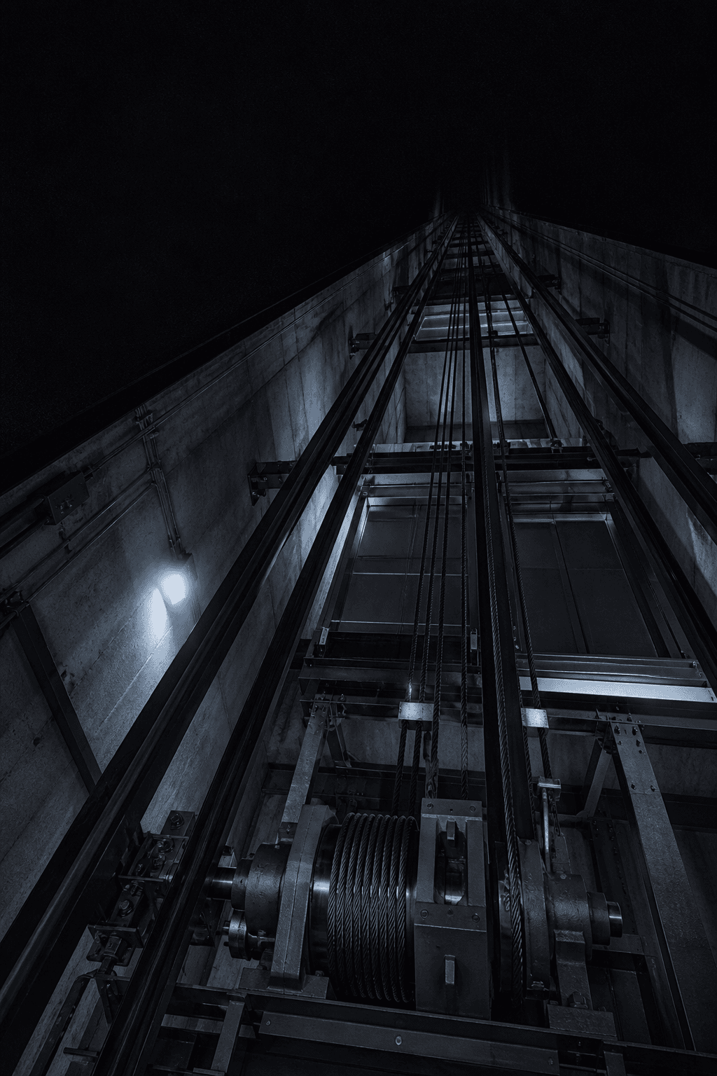 Steel guide rails and cable drums inside a dark elevator hoistway, seen from below